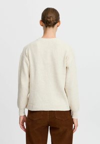 Pull à côtes crème avec manches longues, col rond et coupe décontractée ; associé à un pantalon en velours côtelé marron avec poches à l'arrière.