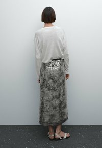 Jupe longue en sequins argentés avec un motif texturé gris, associée à un haut gris clair à manches longues et des sandales blanches. Vue arrière.