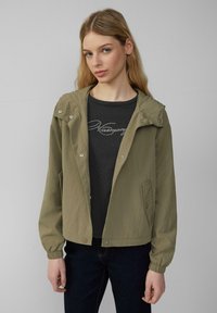 Olijfgroene lichtgewicht jas met capuchon, knopen, elastische manchetten en zijzakken, gedragen over een zwarte getextureerde blouse.