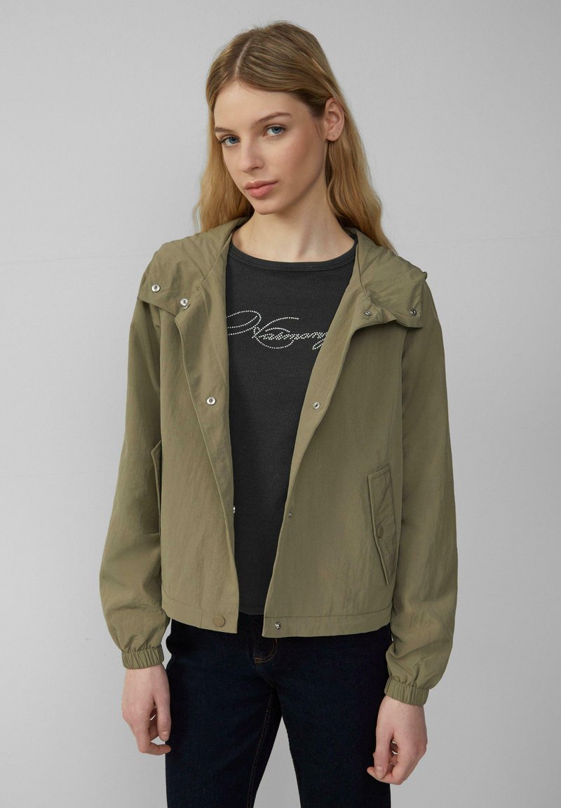 Olijfgroene lichtgewicht jas met capuchon, knopen, elastische manchetten en zijzakken, gedragen over een zwarte getextureerde blouse.