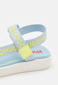 Sandale bleu clair avec des brides imprimées "HUGO" en jaune, une semelle rembourrée et un détail carré jaune sur la bride réglable à la cheville.