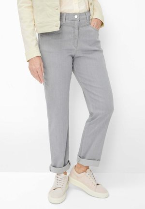 Person wearing light gray cuffed pants, beige sneakers, cream-colored shirt, and light beige jacket, standing with one hand in pocket.