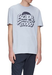 Světle modré bavlněné tričko s velkým tmavě námořnickým grafickým textovým designem, na kterém jsou slova "Escape mood" napsaná zakřiveným tučným písmem.