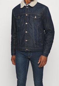 Uomo che indossa una giacca di denim scuro con colletto in pile bianco e jeans di denim scuro abbinati, in piedi davanti a uno sfondo chiaro e semplice.
