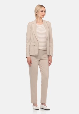 Donna in piedi che indossa un completo beige con pantaloni abbinati, una maglietta beige e bianca a righe e scarpe basse bianche, guarda leggermente di lato.
