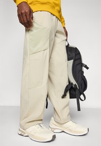 Un pantalon cargo beige clair avec de nombreuses poches, associé à des chaussures de sport blanc cassé. La personne porte un sac à dos noir avec des accents gris.