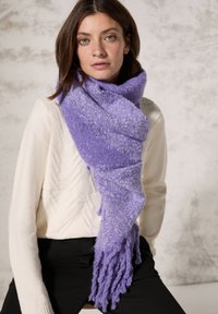Écharpe en tricot violet au toucher doux, présentant un motif dégradé et des bords à franges, joliment enroulée autour du cou, associée à un pull crème.