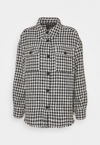 Mustavalkoinen houndstooth-kuvioinen paita-takki, jossa on napitettava etuosa, kaksi rintataskua ja terävä kaulus. Pehmeä, tekstitetty kangas.