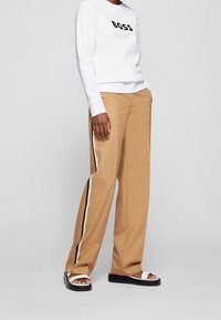 Vit sweatshirt med "BOSS"-logotyp, beige byxor med vida ben och svarta och vita sidostråk, samt svarta sandaler med vita remmar.