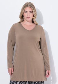 Langärmliges, V-Ausschnitt-Tunika aus hellbraunem Stoff, mit lässiger Passform und glatter Textur, kombiniert mit gemusterten schwarzen Leggings.