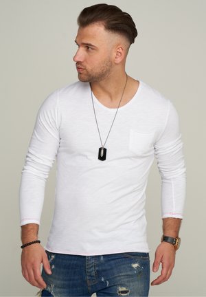 Weißes Langarm-T-Shirt mit einer Tasche, rundem Ausschnitt und roten Nähten, kombiniert mit lässigen blauen Jeans und einer schwarzen Hundemarke als Halskette.