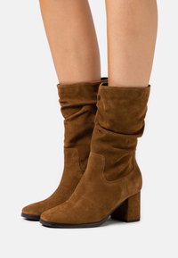 Brown suede mid-calf boots with block heels worn on bare legs, showing soft slouchy texture and rounded toes on a white background.
