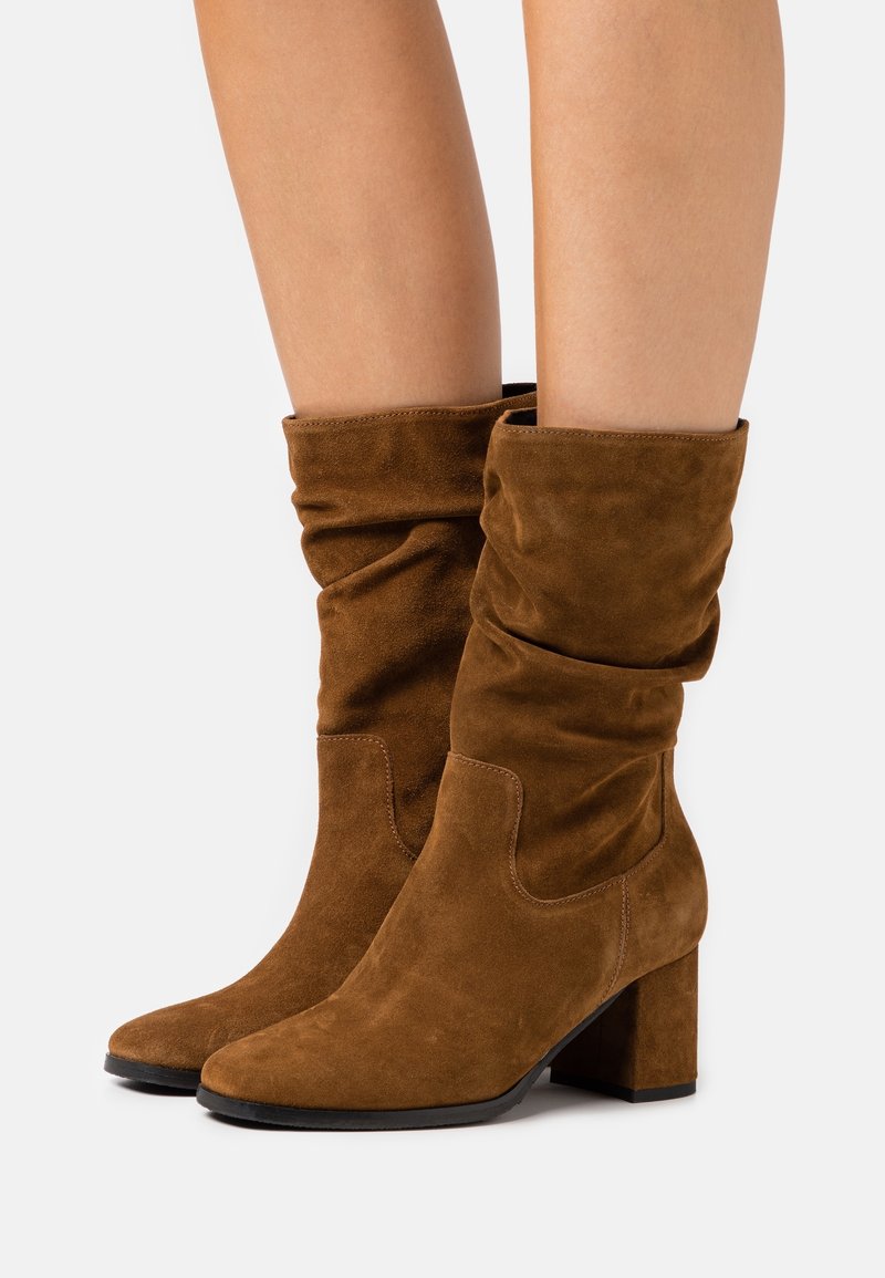 Brown suede mid-calf boots with block heels worn on bare legs, showing soft slouchy texture and rounded toes on a white background.