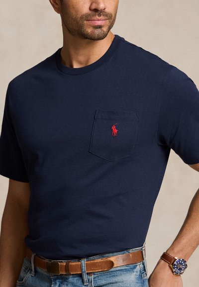 Mann iført navyblå kortermet T-skjorte med rødt logo på brystlommen, brunt skinnbelte, blå jeans og armbåndsur med blå urskive.