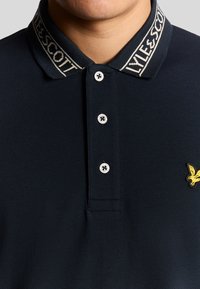 Polo shirt blu navy con colletto a tessuto con "LYLE & SCOTT", tre bottoni argento e logo aquila gialla sul petto.