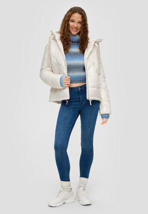 Doudoune couleur crème clair, avec capuche, fermeture éclair à l'avant et poignets élastiques. Portée par-dessus un pull court à rayures bleues et un jean bleu.