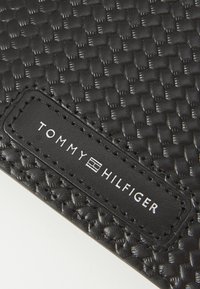 Black woven material with a smooth texture. Features a rectangular leather label with "TOMMY HILFIGER" in white text. No additional patterns.