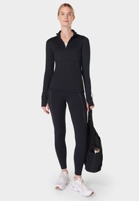 Vrouw in zwarte sportoutfit, met een zwarte sporttas en een waterfles, draagt witte sneakers tegen een lichte achtergrond.