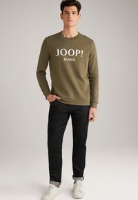 Felpa verde oliva con testo bianco "JOOP! JEANS". Presenta maniche lunghe e polsini a coste, abbinata a jeans scuri e sneakers bianche.