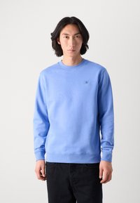 Felpa azzurro chiaro con collo rotondo e polsini a costine; presenta un piccolo logo quadrato sul lato sinistro del petto; abbinata a pantaloni scuri.