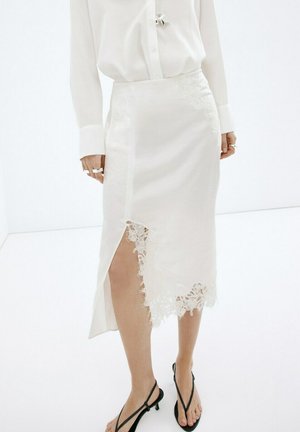 Mujer vistiendo una falda midi de satén blanca con ribete de encaje y abertura, combinada con una blusa blanca y tacones bajos negros con tiras.