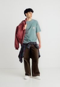 Νεαρός άνδρας με casual εμφάνιση, φοράει μπλουζάκι σε τιρκουάζ χρώμα, καφέ παντελόνι, καρό πουκάμισο δεμένο γύρω από τη μέση, λευκά παπούτσια και κρατάει ένα κόκκινο μπουφάν στον ώμο.