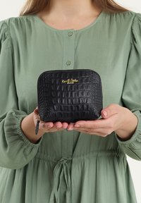 Portafoglio nero con texture a crociera e logo dorato, chiusura a zip e un piccolo tirante, tenuto in mano sopra una blusa verde.