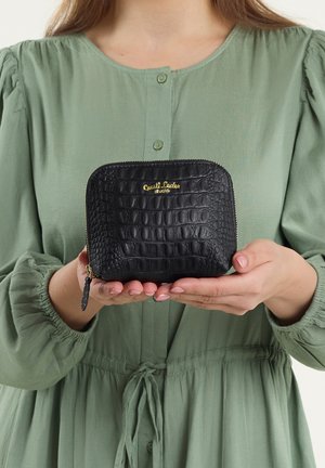 Schwarze Geldbörse mit Krokodilprägung, goldfarbenem Logo, Reißverschluss und kleinem Zipper, in den Händen gehalten vor einer grünen Bluse.