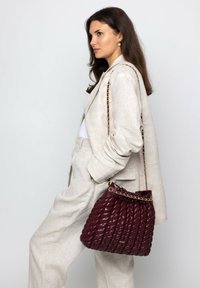 Frau in beigem Anzug mit Händen in den Taschen, die eine gesteppte dunkelrote Lederhandtasche mit goldener Kettenriemen auf der Schulter trägt.