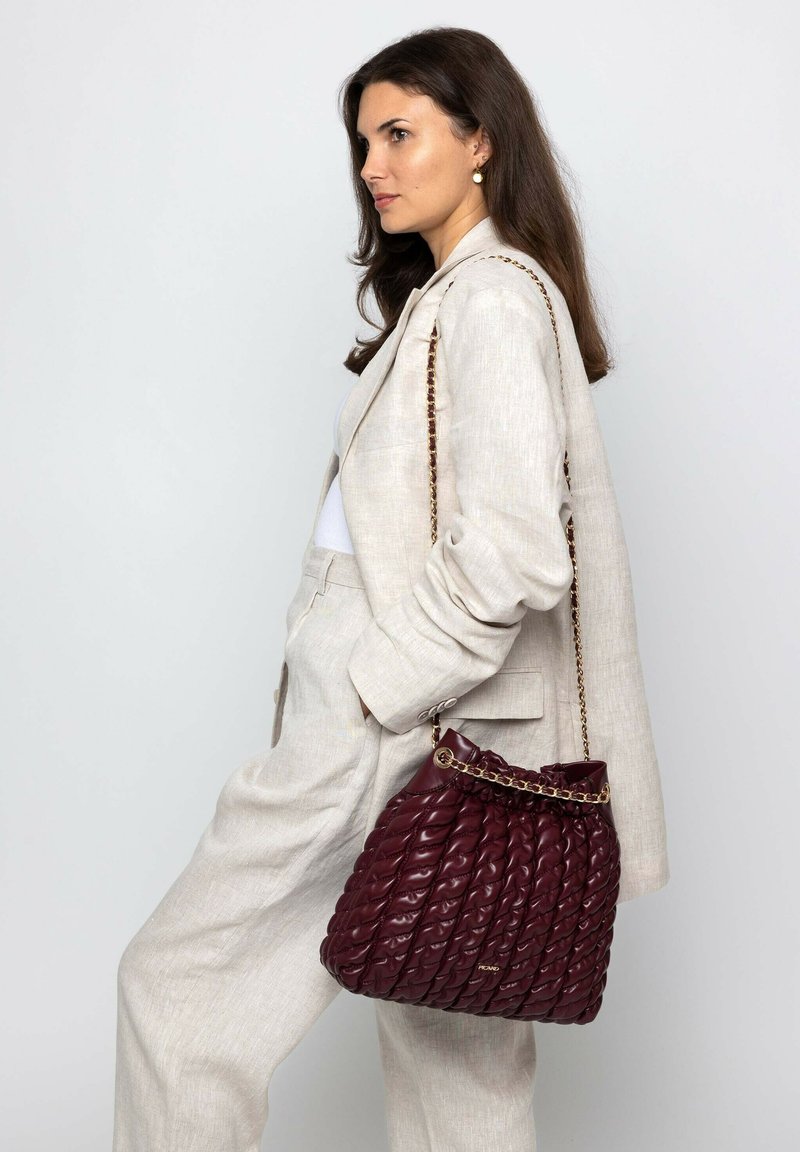 Frau in beigem Anzug mit Händen in den Taschen, die eine gesteppte dunkelrote Lederhandtasche mit goldener Kettenriemen auf der Schulter trägt.