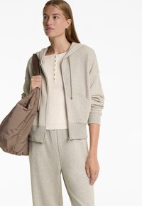 Lichtgrijze ritssluiting hoodie met een voorzak en geribde manchetten, gelaagd over een beige henley-top, gecombineerd met bijpassende sweatpants en een bruine tas.