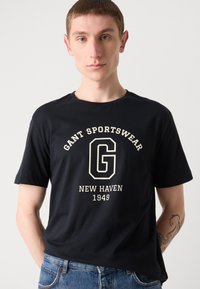 Sort bomulds T-shirt med frontlogo tryk i cremefarve med "GANT SPORTSWEAR" og "NEW HAVEN 1949." Korte ærmer og rund halsudskæring.