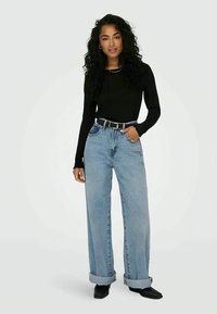 Black long-sleeve top paired with light blue wide-leg jeans, rolled cuffs, and a black belt. Model stands on a plain background.