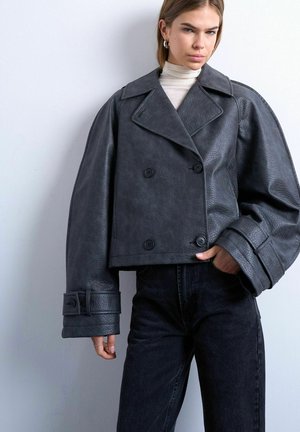 Ung kvinde iført sort, dobbeltradet læderjakke i store størrelser og sorte jeans, stående op ad en lysgrå væg.