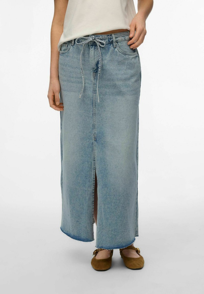 Vrouw draagt een lichtblauwe denim maxi-rok met een split aan de voorkant en een taille met koord, gecombineerd met bruine platte schoenen en een witte top.