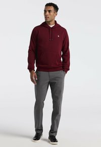 Bordeaux hoodie met een voorzak en geborduurd logo, gecombineerd met grijze broek en zwarte sneakers. Soepele textuur en casual ontwerp.