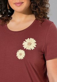 Camisa corta de manga en burdeos con dos margaritas bordadas en crema en la parte superior izquierda, con un cuello redondo y una textura suave.