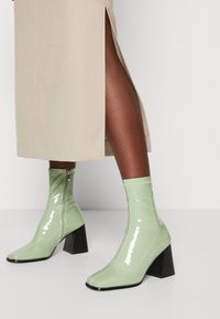 Gröna lackläder ankelskor med fyrkantig tå, en kraftig svart klack och detalj med sidogänga, i kombination med en beige kjol.
