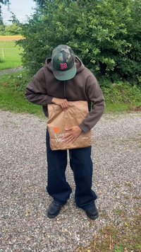 Hnědý papírový sáček s textem "Miluj mě, noste mě" bílou barvou, držený osobou v šedé mikině s kapucí a zelené maskáčové čepici, stojící na štěrku.