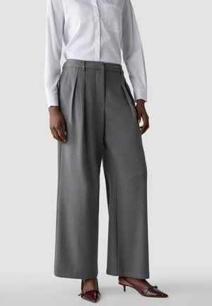 Model trägt hoch taillierte, weit geschnittene graue Hosen kombiniert mit einem weißen Langarmhemd und burgunderroten spitzen Kitten Heels.