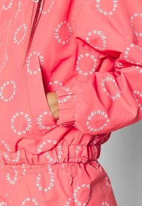 Rosa jacka med upprepat vitt "adidas"-varumärke och logo i ett cirkulärt mönster, elastiska ärmslut och en framficka. Lätt material.