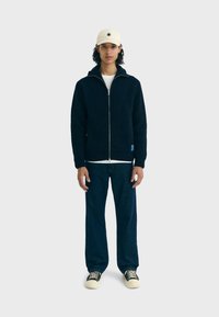 Gebreide marineblauwe zip-up jas met een capuchon, met een geribbelde textuur en V-vormige ontwerpelementen, gecombineerd met donkere denimspijkerbroeken en beige sneakers.