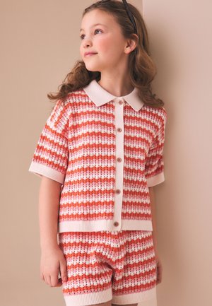 Ung pige iført en lyserød og orange stribet strikket kortærmet skjorte med knapper og matchende shorts, stående mod en beige væg.