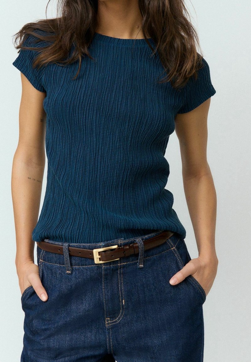 Femme portant un haut bleu foncé texturé à manches courtes, rentré dans un jean bleu taille haute avec une ceinture en cuir marron, les mains dans les poches.