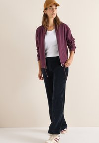 Giacca zip-up bordeaux, t-shirt bianca, pantaloni in corduroy neri, cappellino marrone e sneakers beige con strisce bordeaux. Tessuto liscio con vestibilità rilassata.