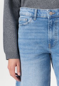 Lys blå denimbukser har høy midje, fem lommer og en enkelt metallknapp øverst. Stoffet har en slitt finish.