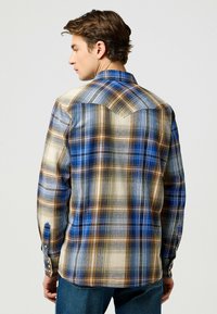 Camisa de cuadros en tonos azul, beige y marrón. Presenta un diseño con yugo, puños abotonados y una textura de tela suave.