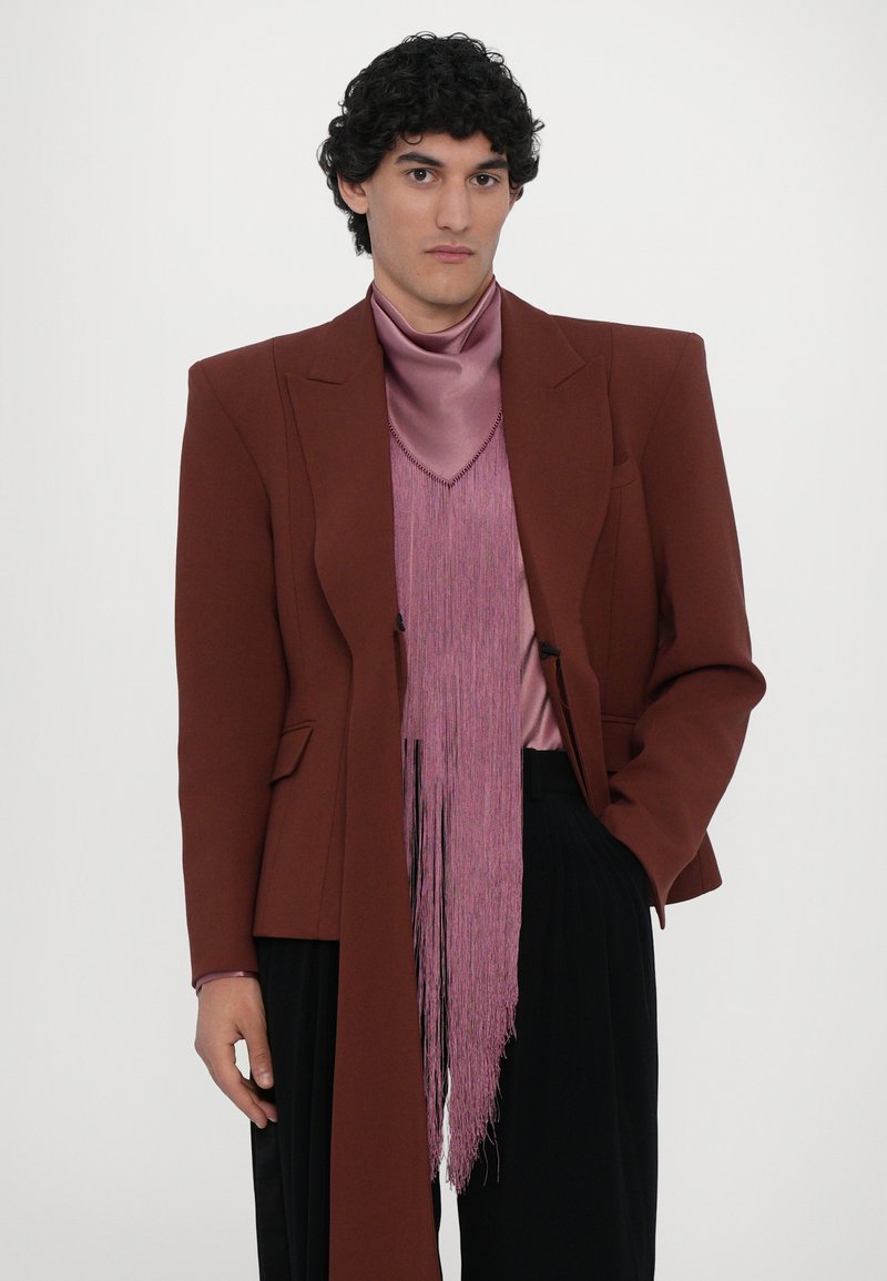 Brauner overgroßer Blazer mit definierten Schultern über einem rosa Satinrollkragenpullover und einer fransigen Weste. Schwarze plissierte Hose rundet das Outfit ab.