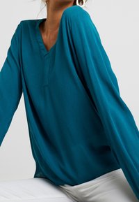 Blouse à manches longues couleur sarcelle avec un col en V, réalisée en tissu texturé, dotée d'une coupe décontractée et de manches légèrement fluides, associée à un pantalon blanc.