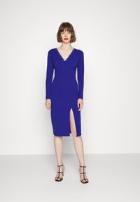 Vestido azul de manga longa com decote em V profundo e acabamento em renda, apresentando uma fenda até a coxa e uma silhueta ajustada. Sandálias de salto preto com tiras.